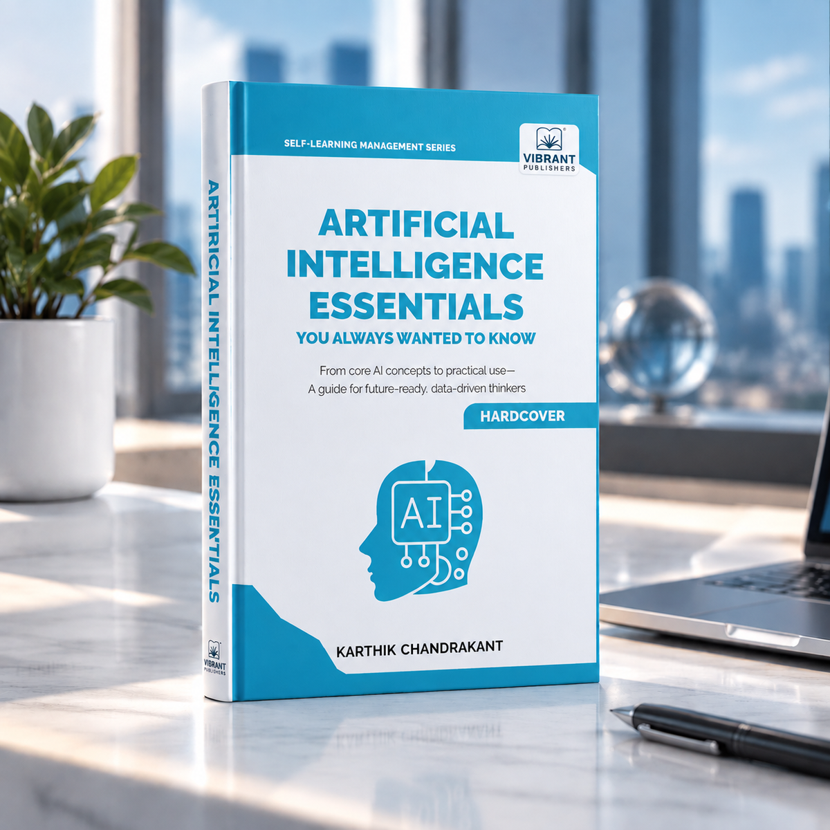 A hardcover book titled 'Artificial Intelligence Essentials' by Karthik Chandrakant, standing upright on a desk with a laptop, pen, and plant in a bright office setting.