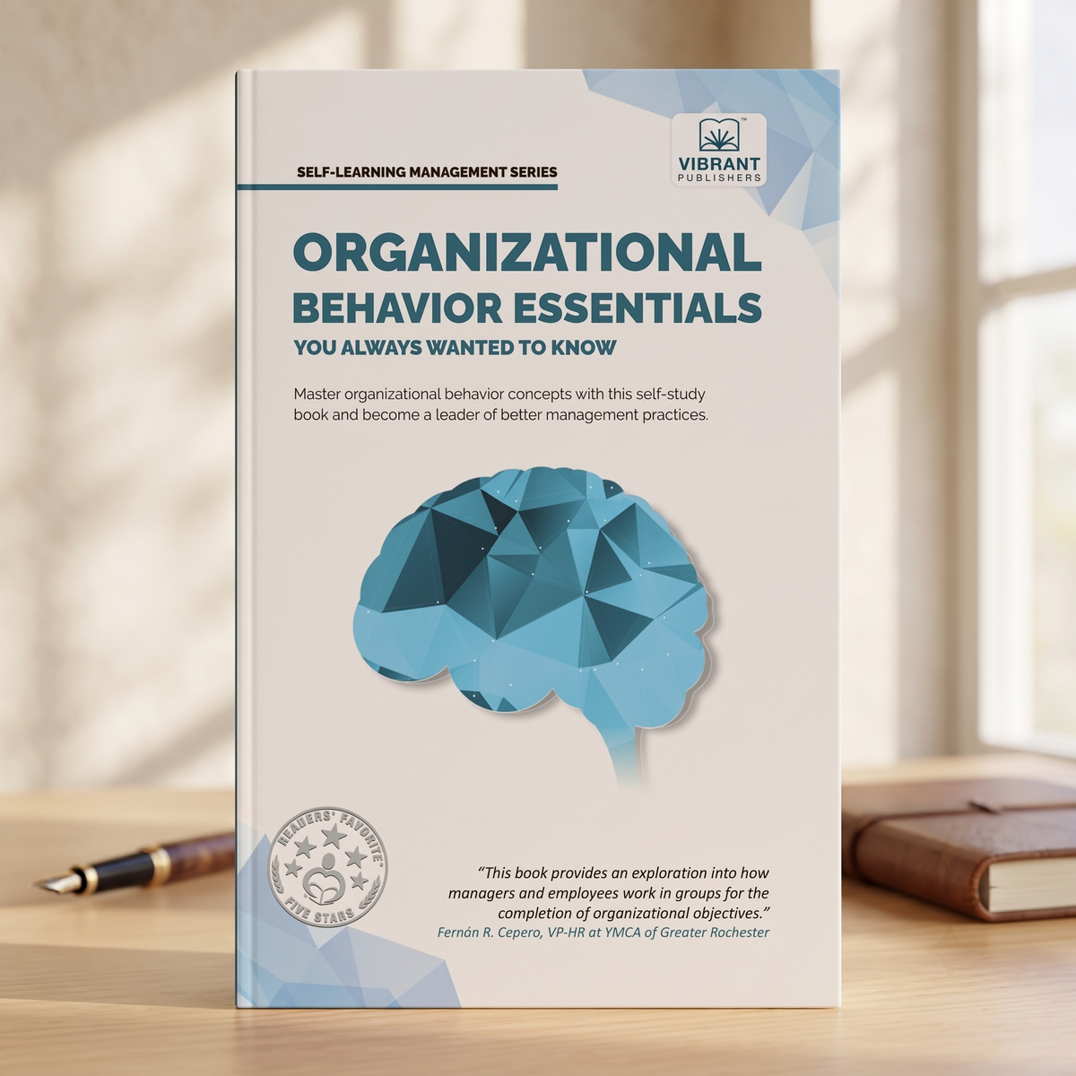 A book titled "Organizational Behavior Essentials" from Vibrant Publishing stands upright on a wooden desk. The cover is white with blue accents, featuring a geometric brain illustration and a blurred office background with a pen and notebook.