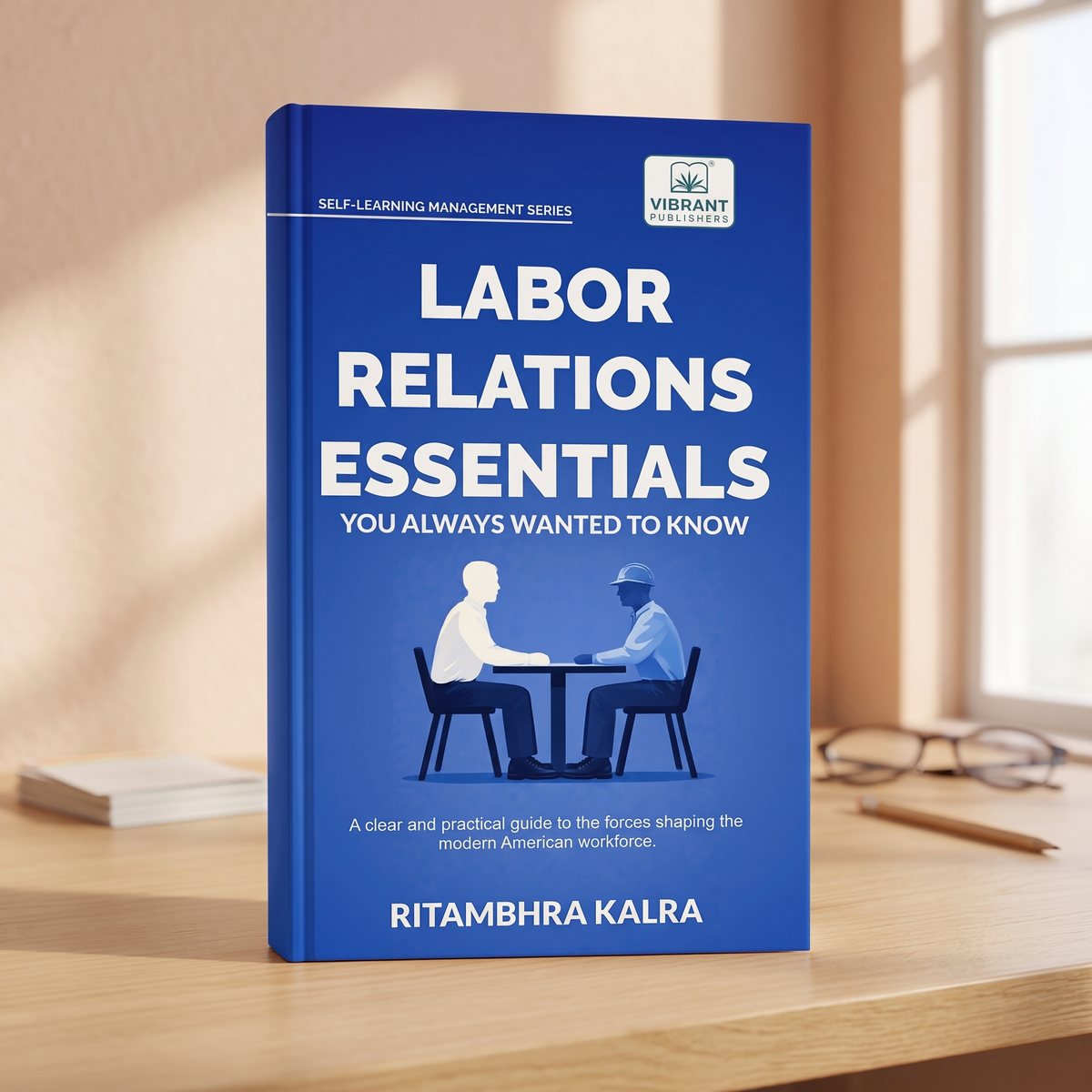 A blue book titled "Labor Relations Essentials: You Always Wanted to Know" by Ritambhra Kalra, standing upright on a wooden desk with warm sunlight and a blurred office background.